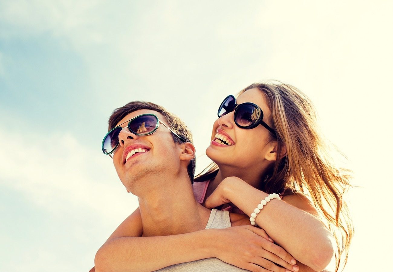 A young couple wearing sunglasses smiles joyfully; the woman is giving the man a piggyback ride under a bright, sunny sky.