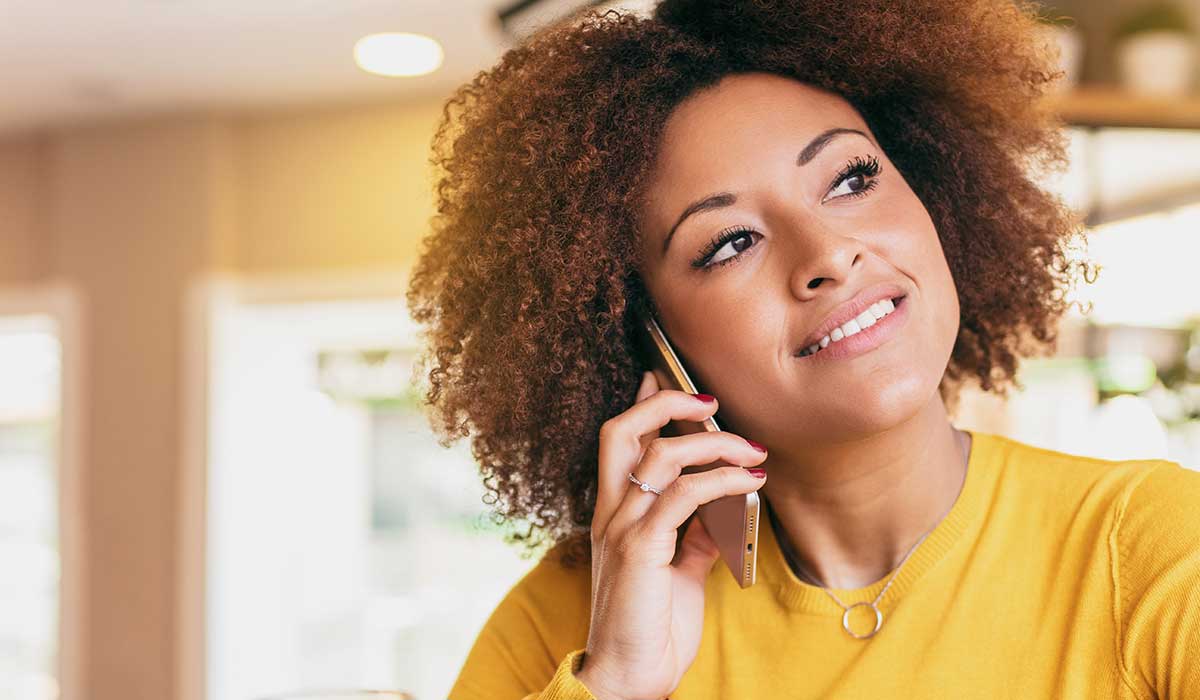 A woman with curly hair wearing a yellow sweater smiles while talking on a smartphone indoors, with soft natural light in the background.