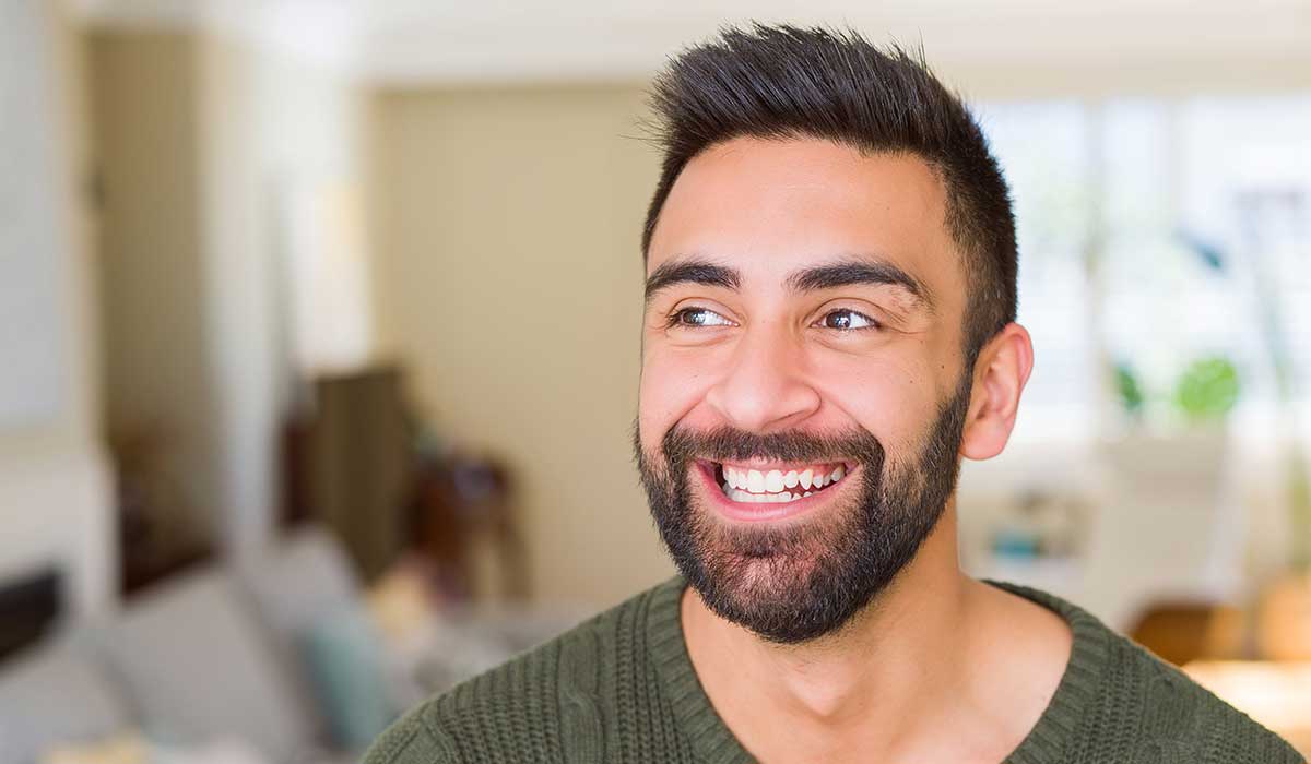A man with short dark hair and a beard smiles while looking slightly to the side, wearing a green sweater. The background is a brightly lit, blurred indoor setting.