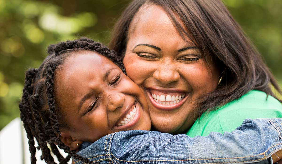 A woman and a young girl, both smiling with their eyes closed, hug each other tightly outdoors. The background is blurred with greenery, highlighting their joyful expressions.