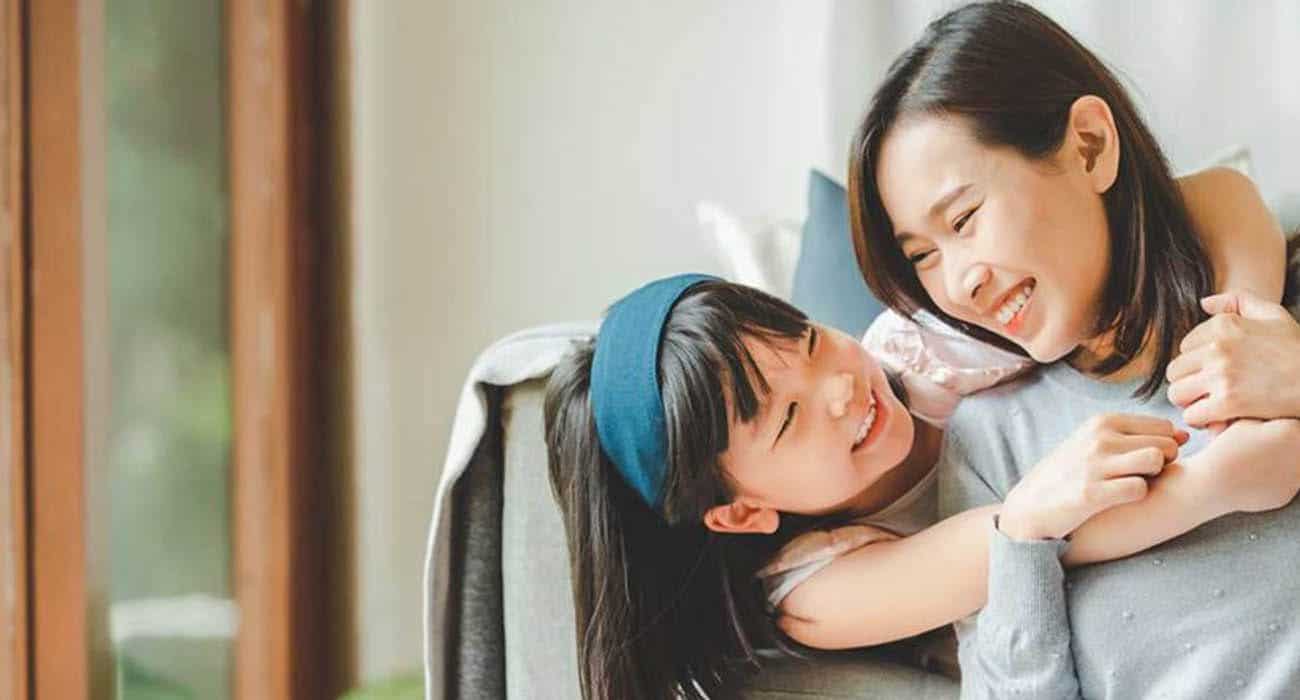 A young girl hugs a smiling woman from behind as they sit on a couch, both looking at each other happily in a bright, cozy living room.